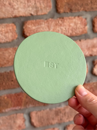 A hand holding a round hand painted sage green textured leather coaste with the Blank Street Logo in the centre, set against a blurred red brick wall background.