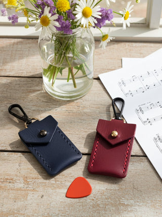 Two small leather coin purses with snap closures and keychain clips are displayed on a rustic wooden tabletop. One pouch is dark navy blue and the other is burgundy red, both with visible contrast stitching and gold-tone snaps.