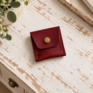 A small burgundy leather card holder or coin purse with a gold snap button, placed on a distressed white wooden surface with a plant in the corner.