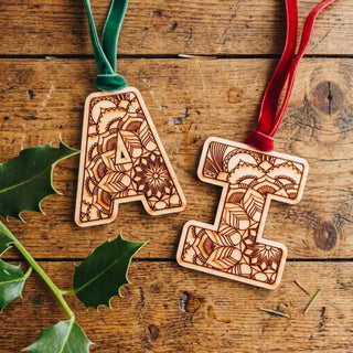 Two wooden letter ornaments, one carved into the shape of 'A' and the other 'I', decorated with intricate floral mandala patterns. The 'A' ornament hangs from a green velvet ribbon, and the 'I' ornament hangs from a red velvet ribbon, displayed on a rustic wooden background with holly leaves.