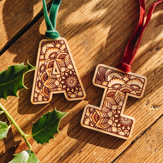 Two laser-engraved wooden letter ornaments, one shaped like the letter A with a green velvet ribbon and one like the letter I with a red velvet ribbon, displayed on a rustic wooden surface with holly leaves.
