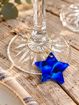 A blue marbled star-shaped glass charm with a gold ring loop, hanging from a clear wine glass stem on a rustic wooden table.