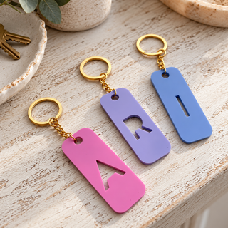 Three rectangular metal keychains with gold key rings are arranged on a light wood surface. Each keychain has a different solid color: pink with a pink letter 'A', lavender with a purple letter 'R', and blue with a blue letter 'I'.
