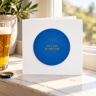 A white square greeting card with a large blue circular cutout in the center and gold text reading 'DAD'S BEER BELONGS HERE', photographed on a sunlit windowsill with a glass of beer and a plant nearby.