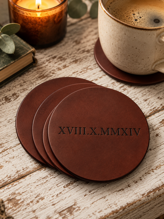 Four round mahogany brown leather coasters stacked on a rustic white-washed wooden table. The front coaster is embossed with the Roman numerals 'XVIII.