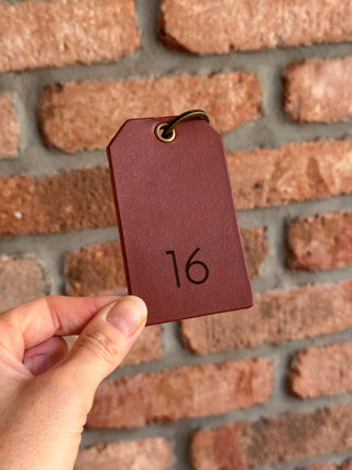 A hand holding a small tan leather tag with a brass grommet and the number 16 engraved on it, set against a rustic brick wall background.