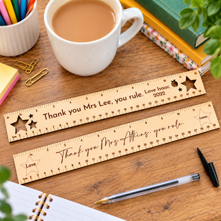 Two personalised wooden ruler desk toppers with engraved text and star cutouts, displayed on a wooden desk alongside a white coffee mug, notebook, pen, paper clips, and books.