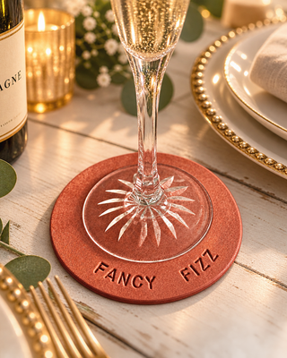 A pink-coloured round coaster engraved with 'FANCY FIZZ' sits on a white wooden table, supporting a champagne flute. The scene is styled with a bottle of champagne, gold candlelight, eucalyptus leaves, and gold-rimmed plates.