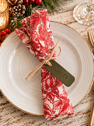 A folded place napkin with a red and cream toile de Jouy floral pattern, tied with a twine bow and a dark olive green tag engraved with the name 'JAMES', presented on a white ceramic plate with gold trim.