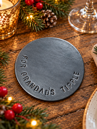 A round dark gray cast-iron coaster engraved with 'FOR GRANDAD'S TIPPLE' in a holiday-themed setting on a wooden table with pine branches, red berries, and a candle.