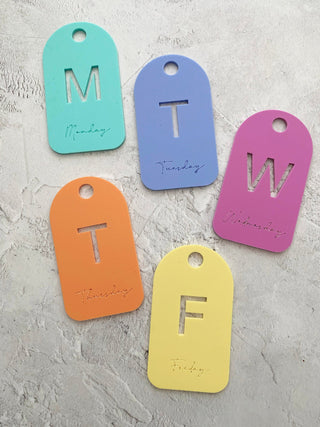Five colourful tags with letters for each day of the week on a textured surface.
