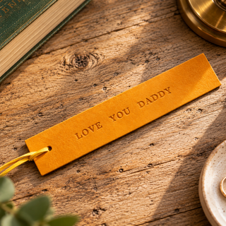 A yellow leather bookmark engraved with 'LOVE YOU DADDY' and attached with a yellow string, resting on a rustic wooden tabletop with styled props.