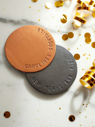 Two round coasters with embossed text on a marble surface with gold confetti and ribbons.