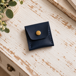 A small navy blue leather card holder or coin pouch with a gold snap button, placed on a distressed white wooden surface with a plant in the corner.