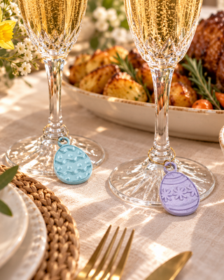 Two decorative egg-shaped drink strainers in light blue and lavender purple, hanging from champagne glasses on a beautifully set dining table with roasted meat, potatoes, and flowers in the background.