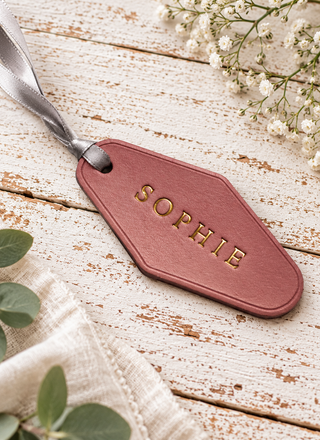 A pink leather luggage tag with the name 'SOPHIE' embossed in gold, resting on a rustic white wooden surface with baby's breath flowers and green leaves.