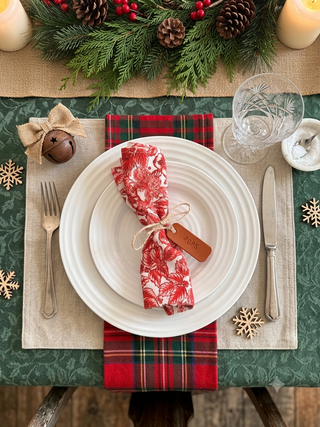 A festive holiday table setting featuring a place setting with a red and white patterned napkin tied with twine, white plates, silver cutlery, a green plaid runner, and a green tablecloth decorated with pinecones, evergreen branches, and candles.