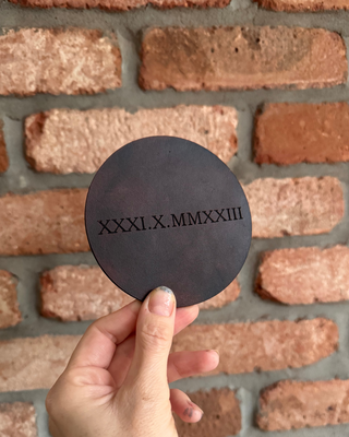 A hand holding a round black leather coaster with the Roman numerals 'XXXI.X.