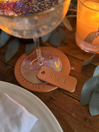 Cocktail glass with a branded coaster on a wooden table with leather wine glass charm stamped with Rosie