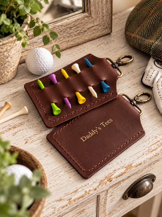 A brown leather golf tees organizer wallet with brass key rings, shown on a rustic wooden table surrounded by golf props including golf balls, tees, a hat, and a mirror.
