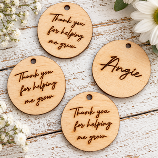 Four round wooden tag charms with hanging holes, three engraved with 'Thank you for helping me grow' and one personalized with 'Angie', arranged on a whitewashed wooden surface with white flowers.