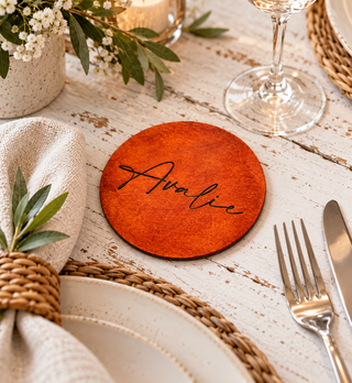 A round wooden place coaster engraved with the name 'Avalie' in black script, styled on a rustic white-washed wooden table with a beige napkin, greenery, a wine glass, and silver cutlery.
