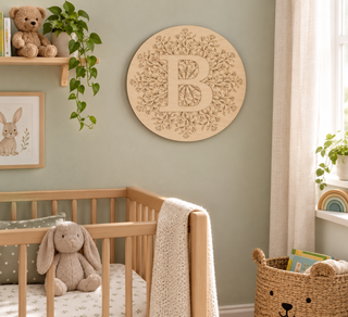 A round wooden wall art piece featuring a carved monogram letter B surrounded by delicate floral scrollwork, displayed in a styled nursery above a wooden crib.