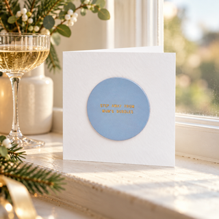 A white square greeting card with a circular light blue center and gold foil text reading 'STEP AWAY FROM BAD TIMES' stands on a sunlit windowsill. A wine glass, greenery, and soft outdoor background are visible around it.