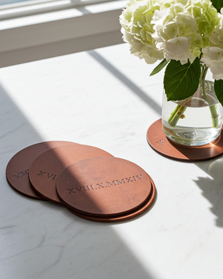 Tan Leather coasters with engraved numbers on a white surface with a vase of white flowers.