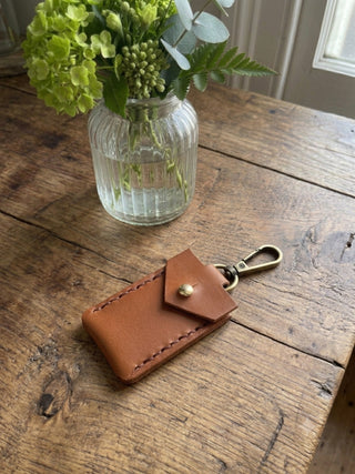 A small tan leather coin holder or wallet with a sam brown fastening and brass keychain clip, placed on a rustic wooden table next to a glass vase with green flowers.
