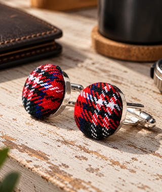 A pair of round cufflinks with red, black, and white tartan plaid fabric inserts and silver-toned metal hardware, displayed on a rustic wooden tabletop with blurred lifestyle props in the background.