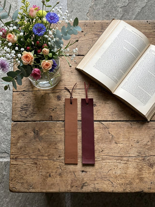Two leather bookmark tags, one tan and one burgundy, displayed on a rustic wooden table next to an open book and a glass vase of flowers.