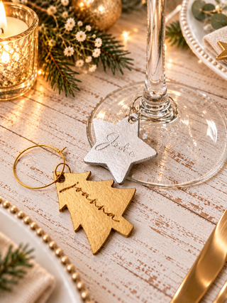 Personalized Christmas tableware featuring a gold wooden tree-shaped tag engraved with 'Jonathan' and a silver star-shaped tag engraved with 'Joy', both attached to a gold wine glass stem on a rustic white wooden table decorated with a candle, pine branches, and fairy lights.