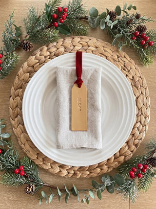 A styled holiday table setting featuring a white ceramic plate on a round woven straw placemat, with a folded cream linen napkin and a personalized wooden tag tied with a red ribbon. The scene is decorated with green pine branches, eucalyptus leaves, and red berries on a light wood tabletop.