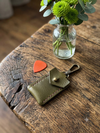 An olive green leather guitar pick holder with a snap button and metal clip, displayed on a rustic wooden table next to an orange guitar pick, with a small glass vase of green flowers in the background.