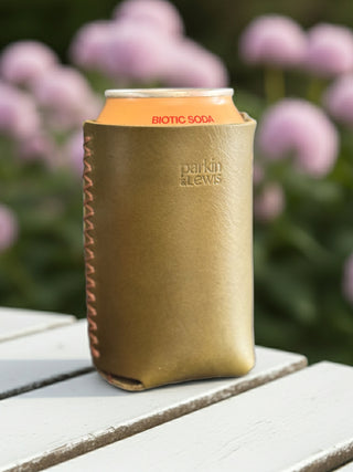Green leather can cooler with 'Biotic Soda' can on a wooden surface with blurred flowers in the background